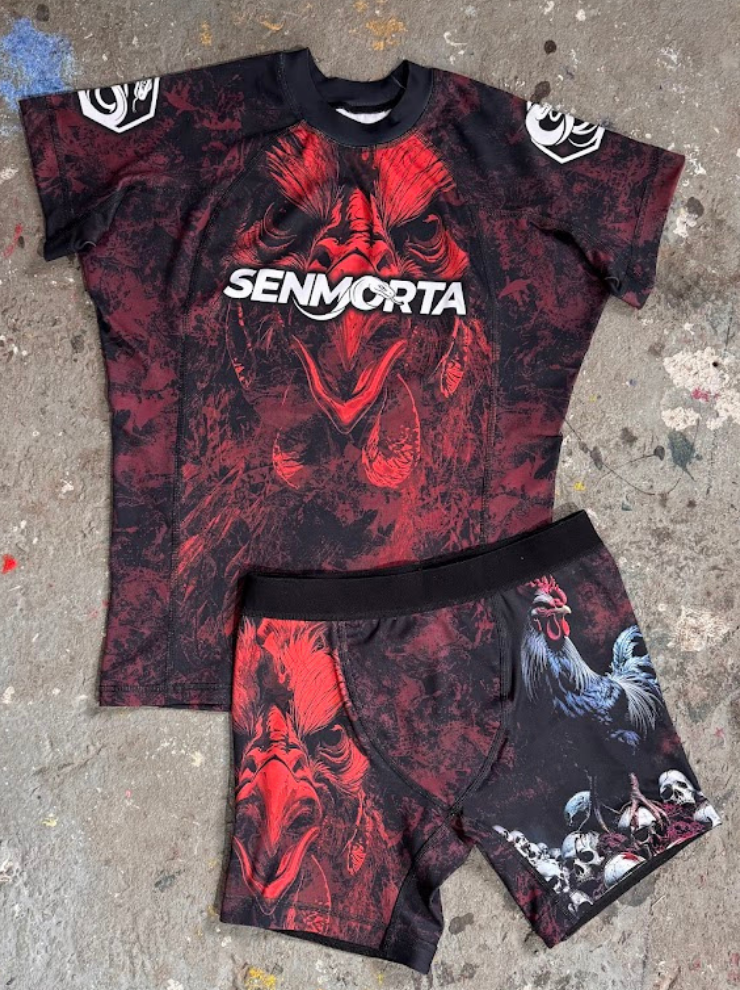 Komplet Bloody Rooster, przedstawia rashguard ze wzorem czerwonego koguta z napisem senmorta  pośrodku,. Niżej są spodenki, prawa nogawka z nadrukowanym czerwonym kogutem takim samym jak na rashguardzie, a lewa nogawka z niebieskim kogutem stojącym na stosie zakrwawionych czaszek