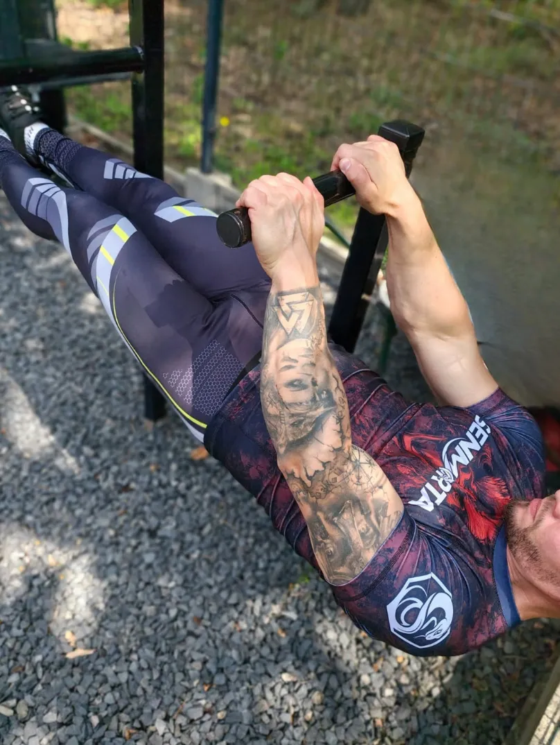 mężczyzna podciągający się na leżąco na drążku, ubrany jest w rashguard Bloody Rooster ze wzorem czerwonego koguta